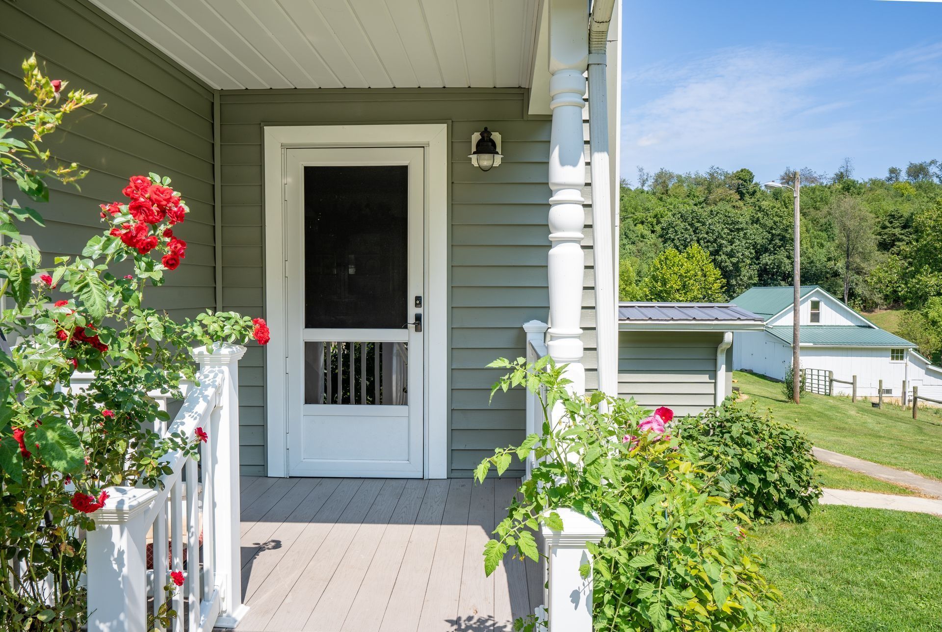 Front porch with white door, green siding, and red flowers overlooking a lake and trees
