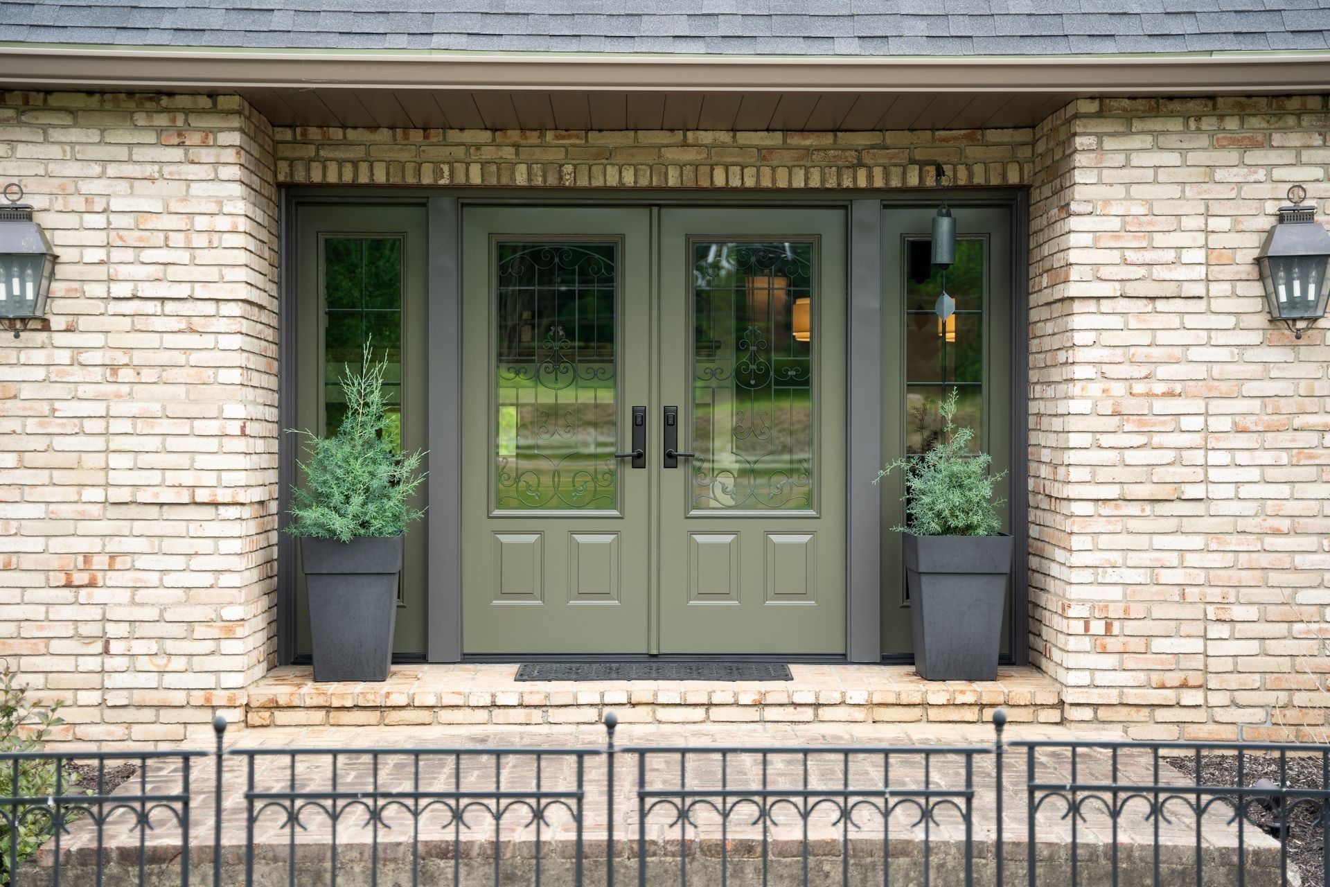 Front entrance with green double doors, sidelights, and potted plants against a light brick facade.