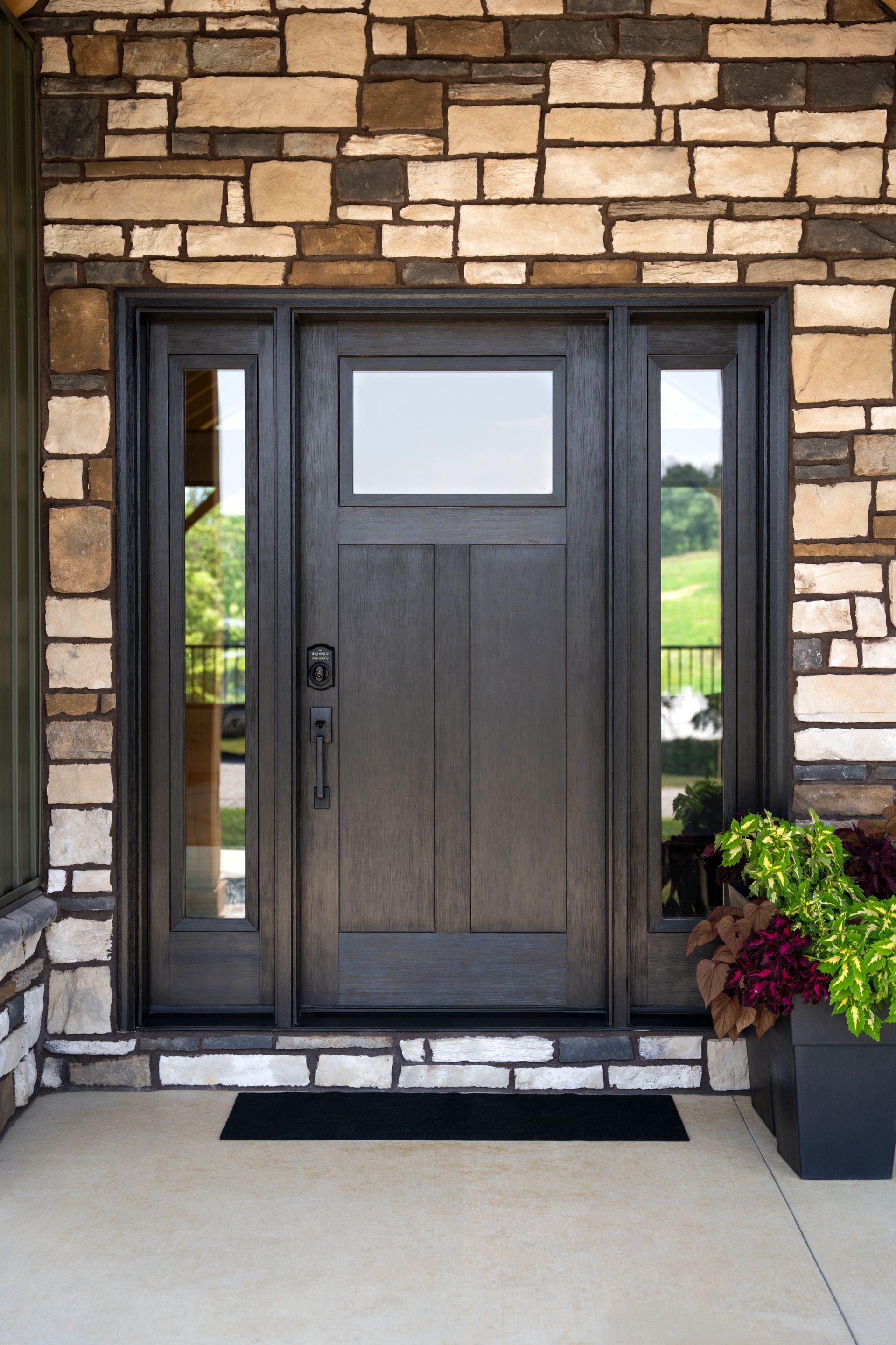 Front entrance with dark wood door and sidelights in a stone wall, with a planter and doormat.