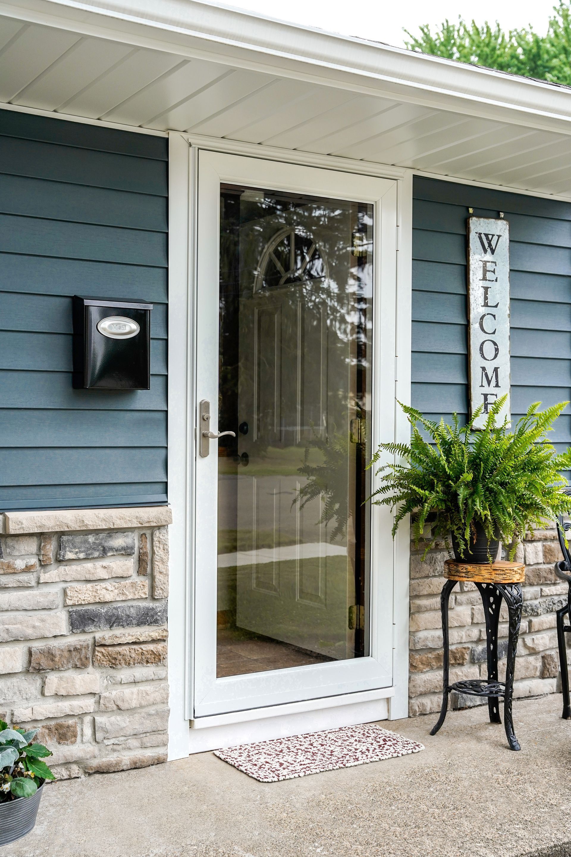 Front porch with white glass storm door, blue siding, welcome sign, and potted plant on a small table.