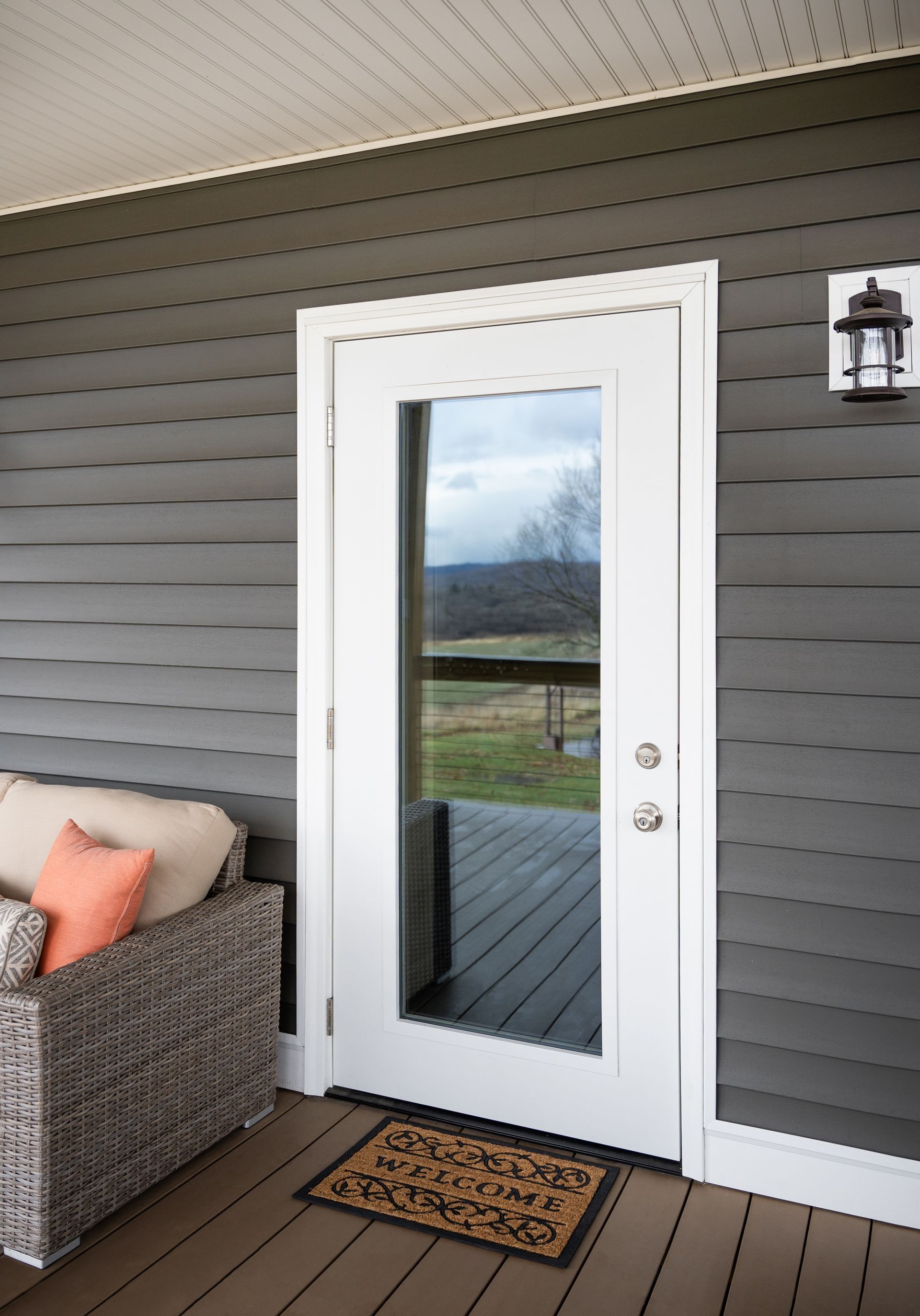Porch with white glass door, dark siding, wicker chair, and doormat overlooking a yard.
