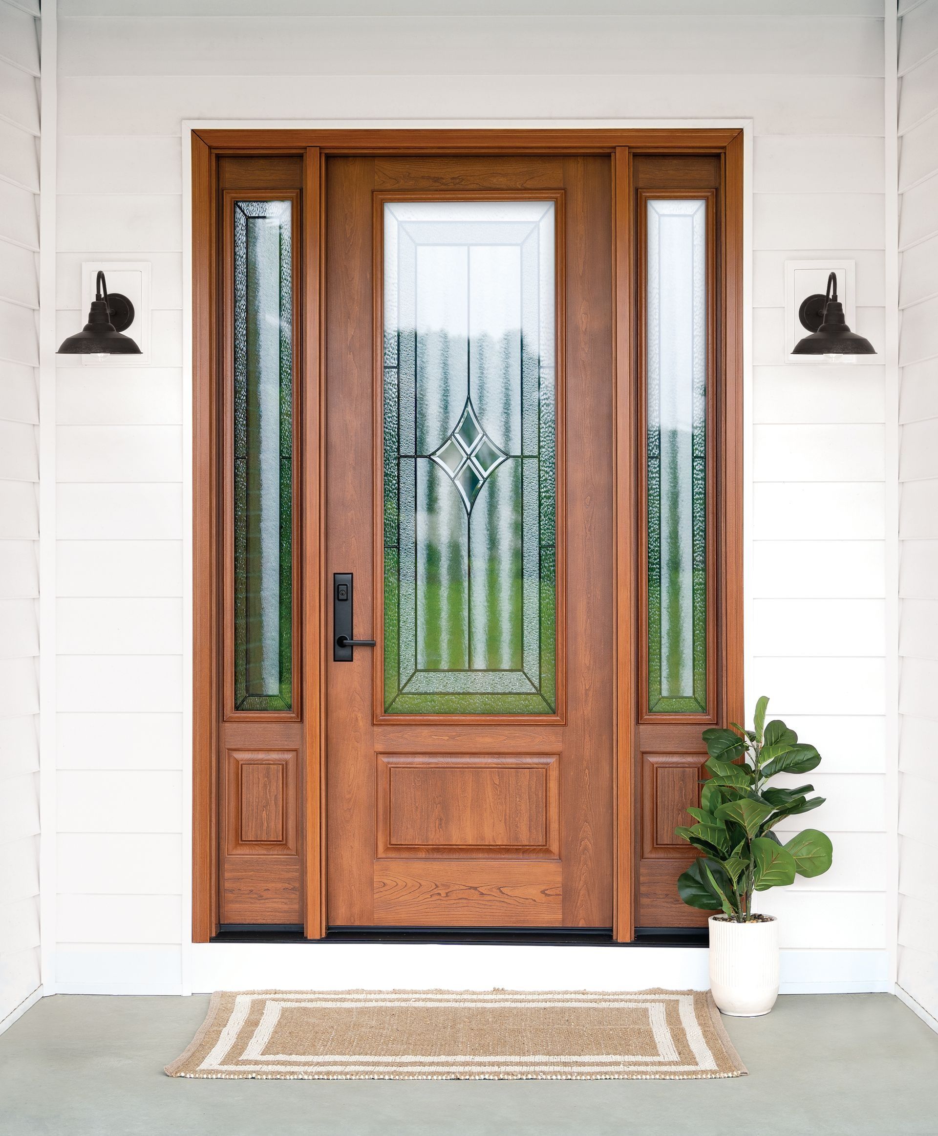 Wooden front door with glass panels, flanked by black sconces, white siding, and a potted plant on the porch.