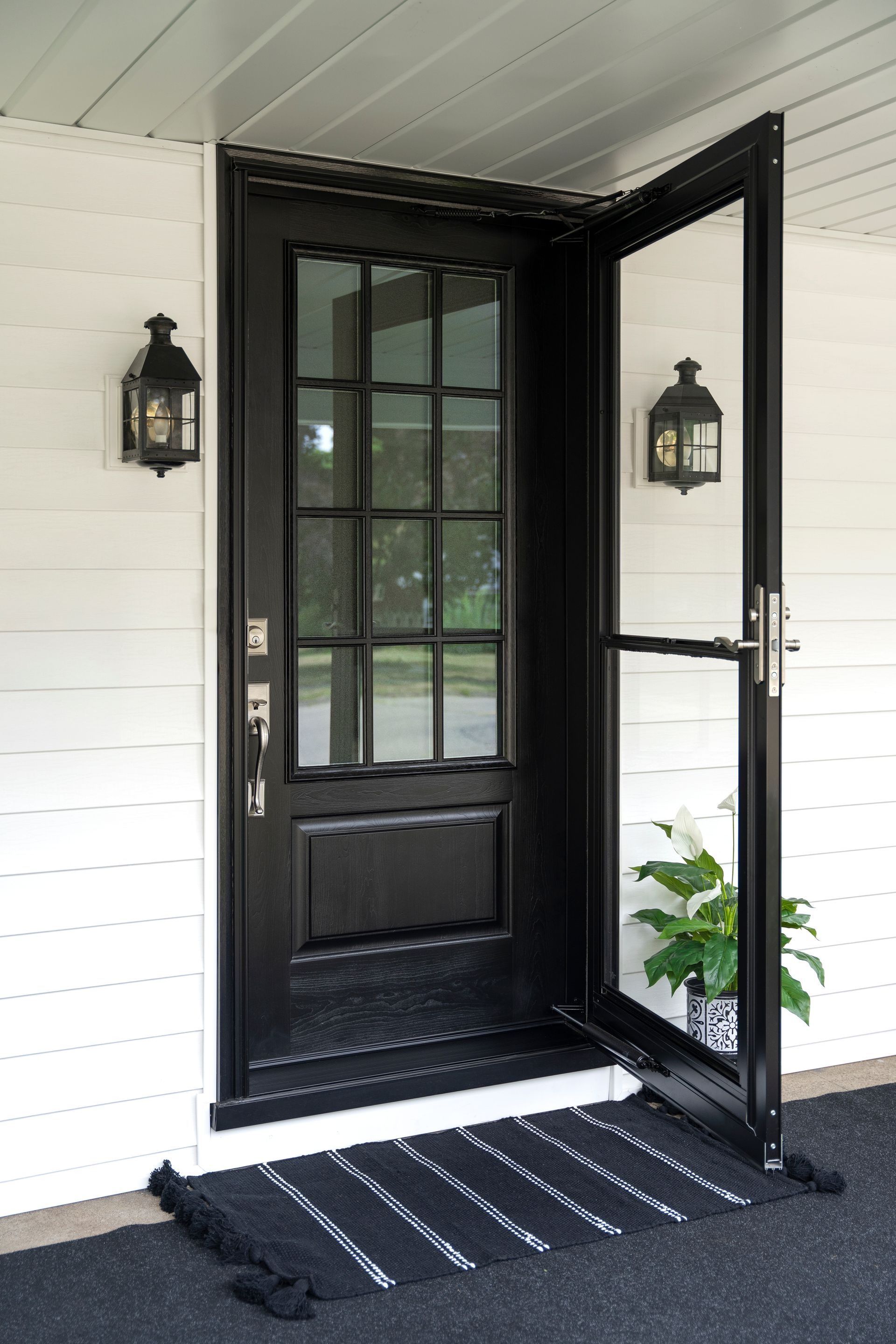 Front porch with open black door, wall lanterns, and striped rug on a white house exterior.