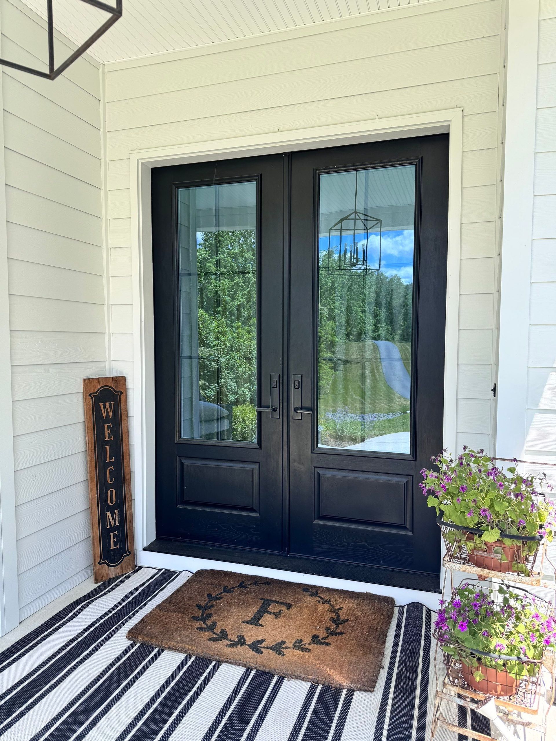 Black double front doors with glass panes, doormat, and potted flowers on a porch.