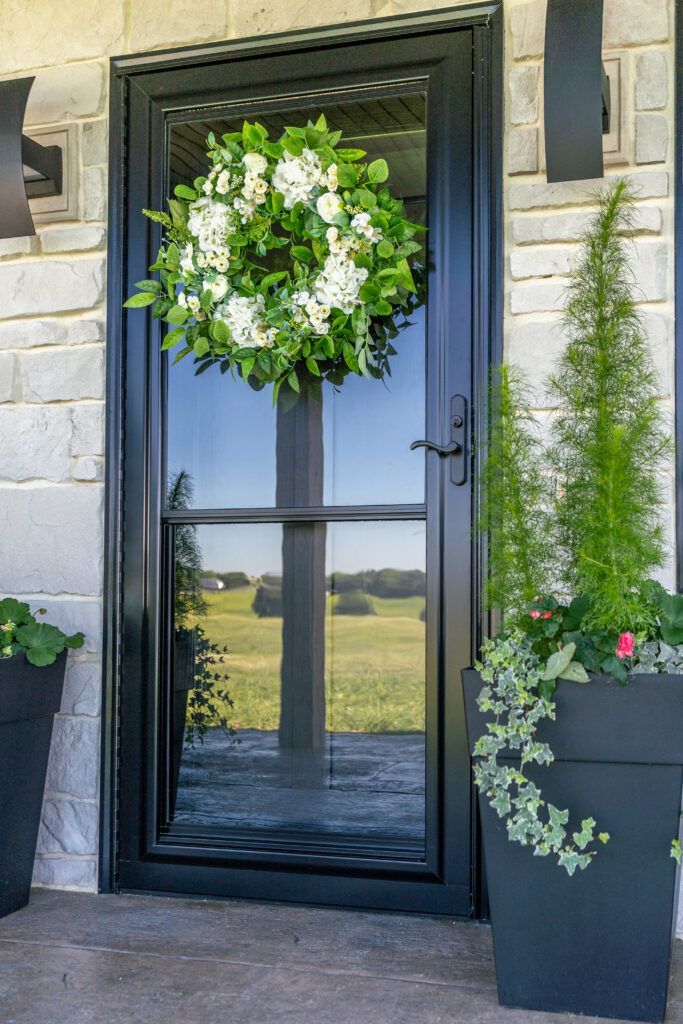 Front door with a green wreath, glass pane reflecting a landscape, and potted plants on each side.