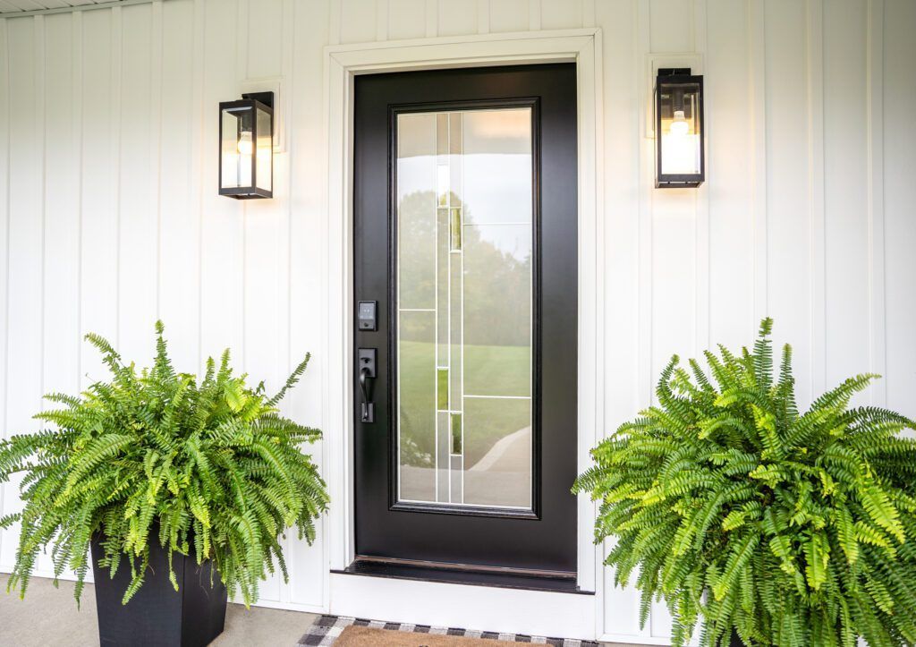Front porch with black front door, wall lanterns, and two large green ferns in planters.