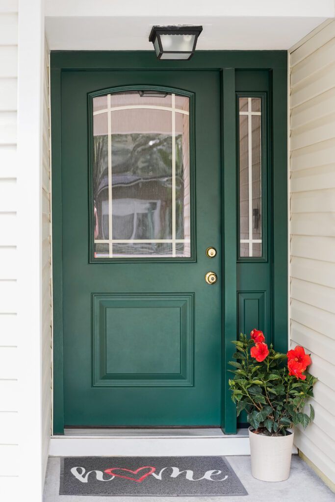 Green front door with glass panels, a welcome mat, and a potted red flower beside it