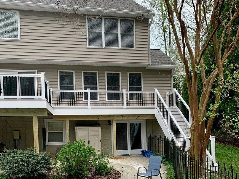 Two-story beige house with a white balcony, staircase, and blue patio chair in a wooded backyard