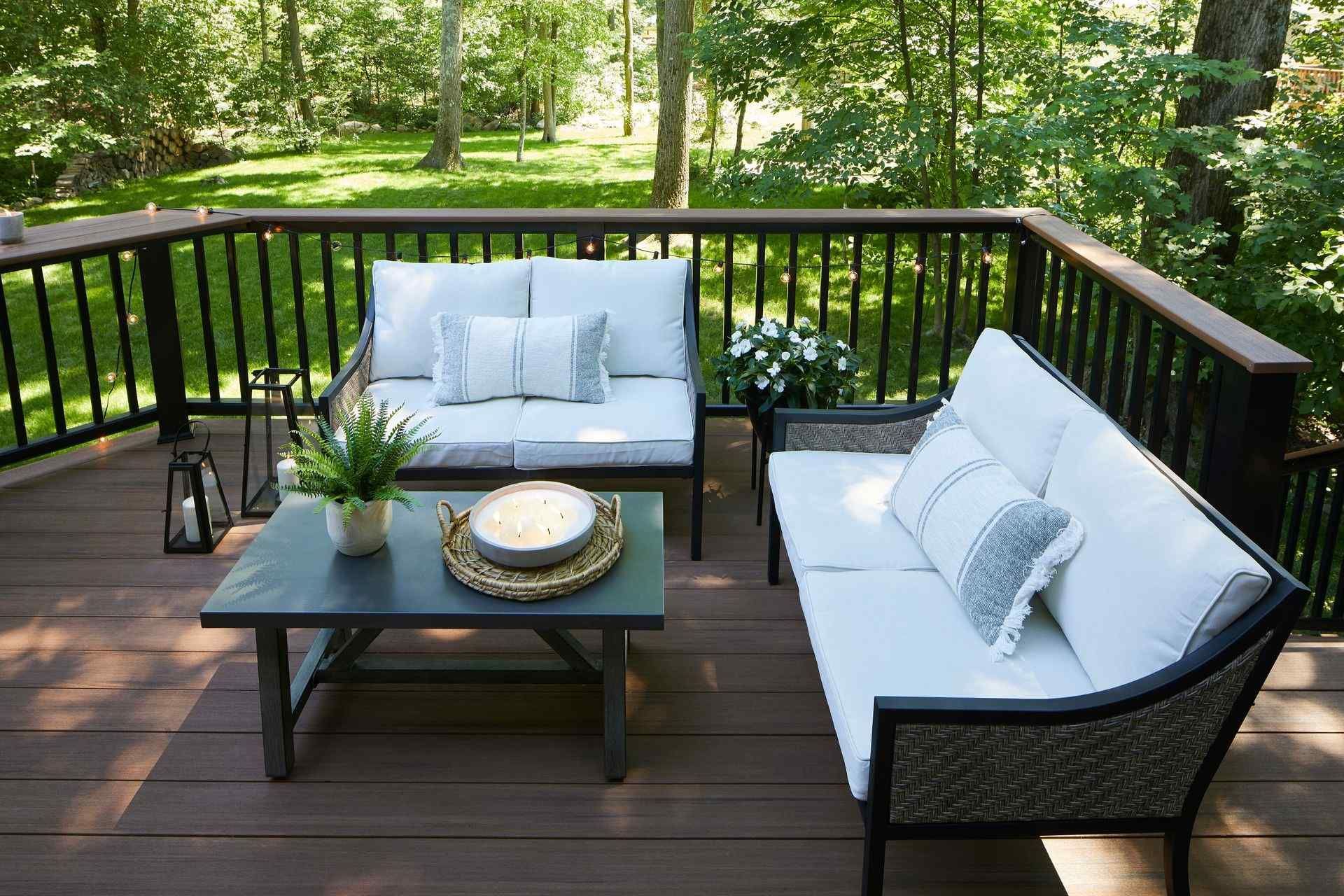 Outdoor wooden deck with white cushioned patio seating, black railing, and a small coffee table amid green trees