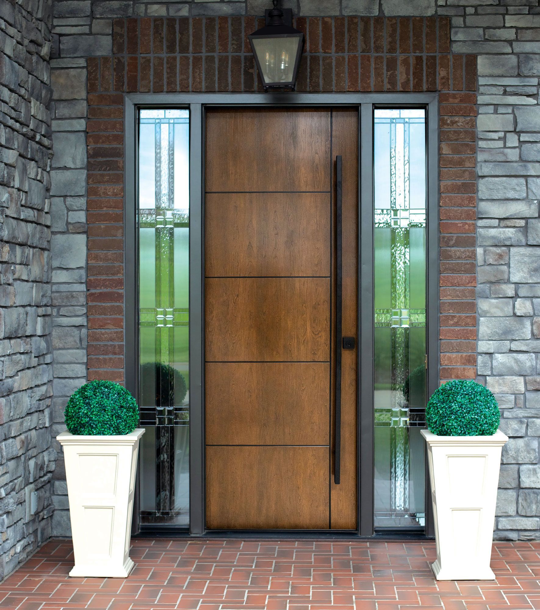 Modern wooden front door with narrow glass panels, flanked by stone walls and two white planters with green shrubs