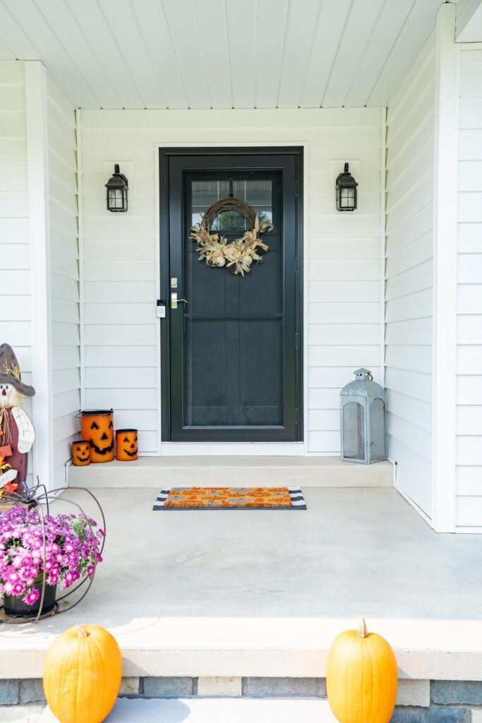 Front porch with black door, fall wreath, white siding, pumpkins, and flowers on the steps.