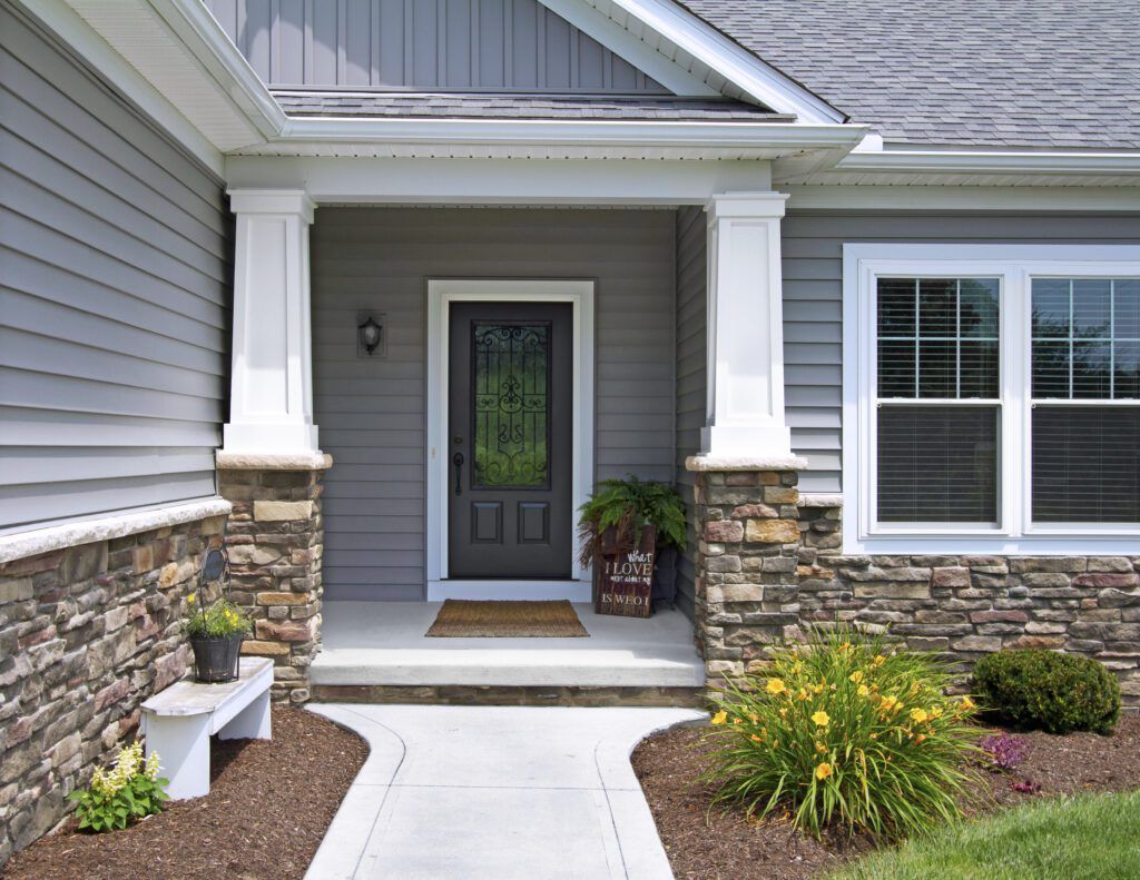 Front porch entrance with gray door, stone siding, white columns, and flower beds