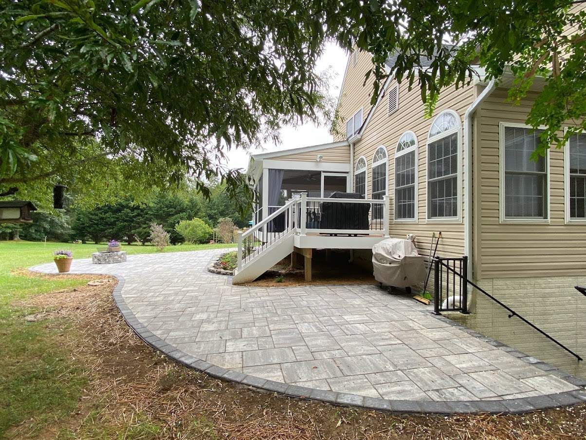 Stone patio wraps around a beige house with a white-railed porch under leafy trees.