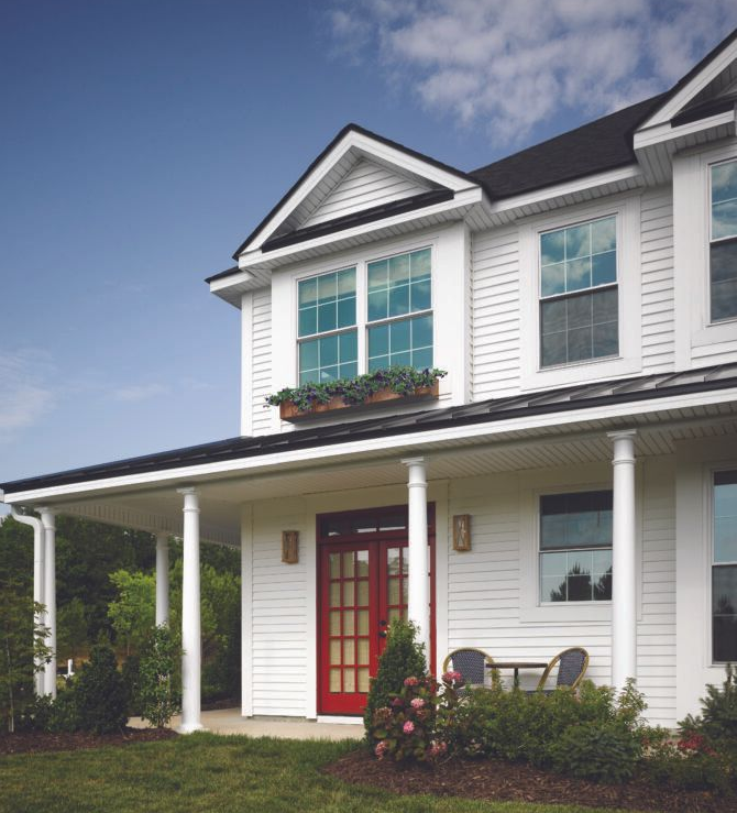 Two-story white house with red front door, green shutters, and a covered porch under a blue sky