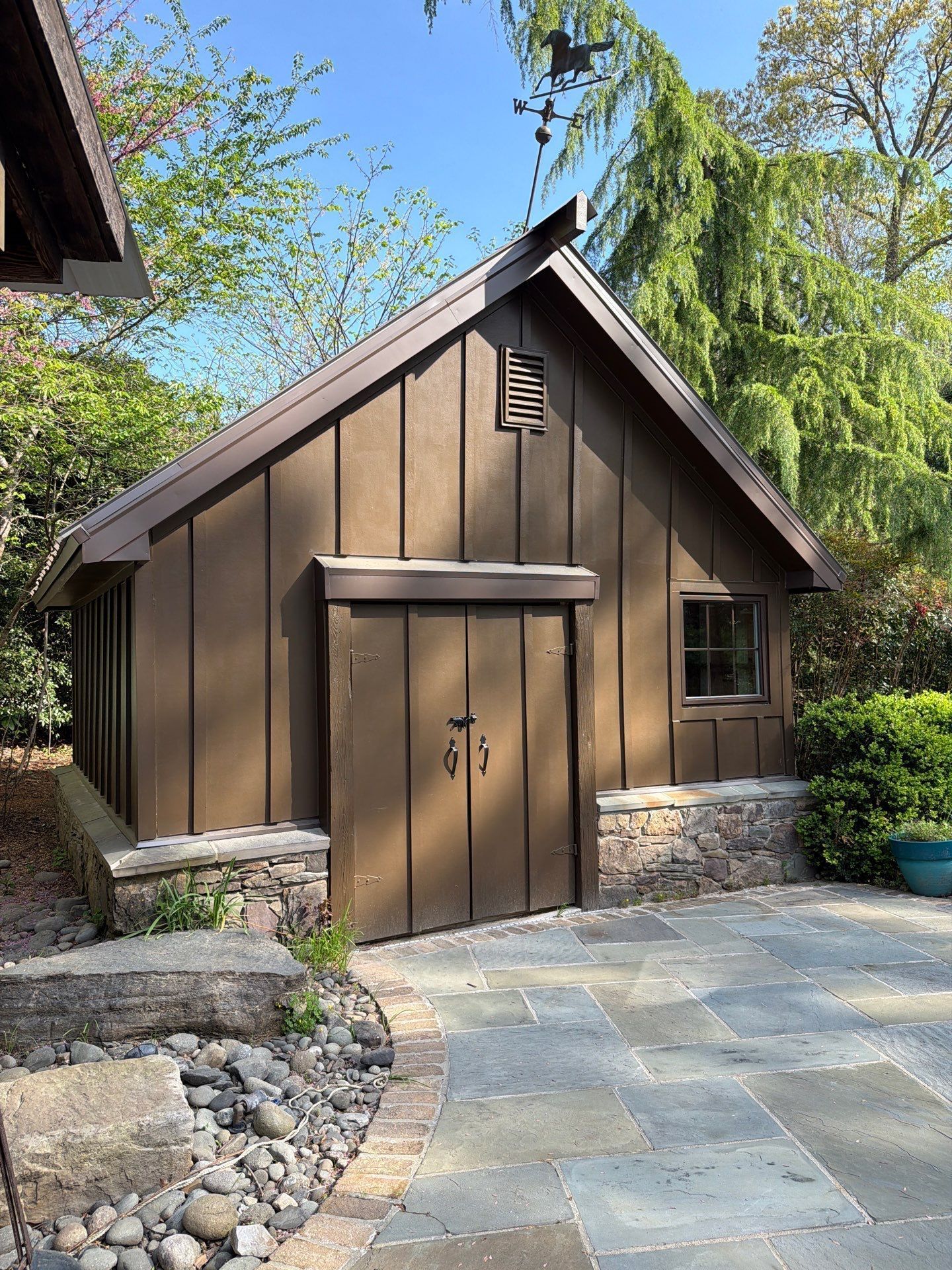 Small rustic brown shed with double doors, stone trim, and a window in a landscaped garden.