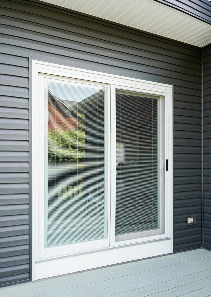 White sliding glass patio door with blinds on a dark gray house exterior and light gray deck.