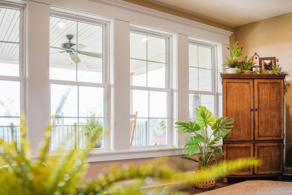 Sunlit living room with large windows, a wooden cabinet, and leafy houseplants.