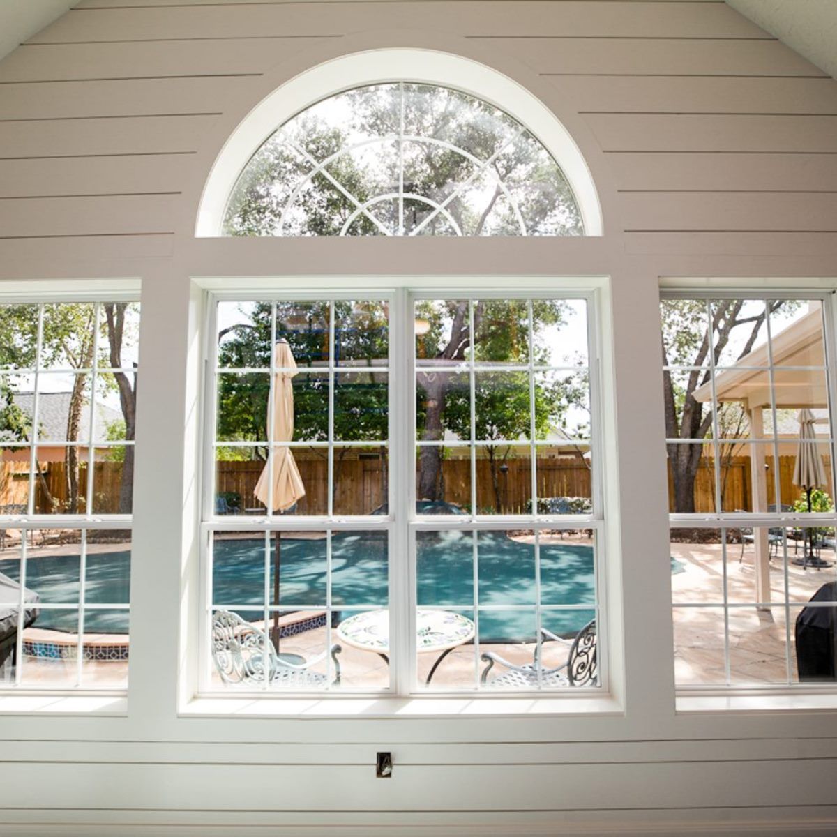 Bright sunroom windows overlook a backyard pool and patio with white chairs and umbrella