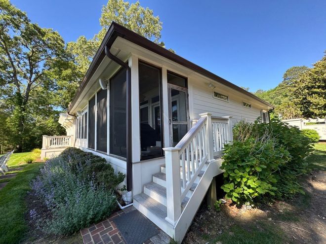 White house porch with screened windows, front steps, and lush green landscaping on a sunny day.