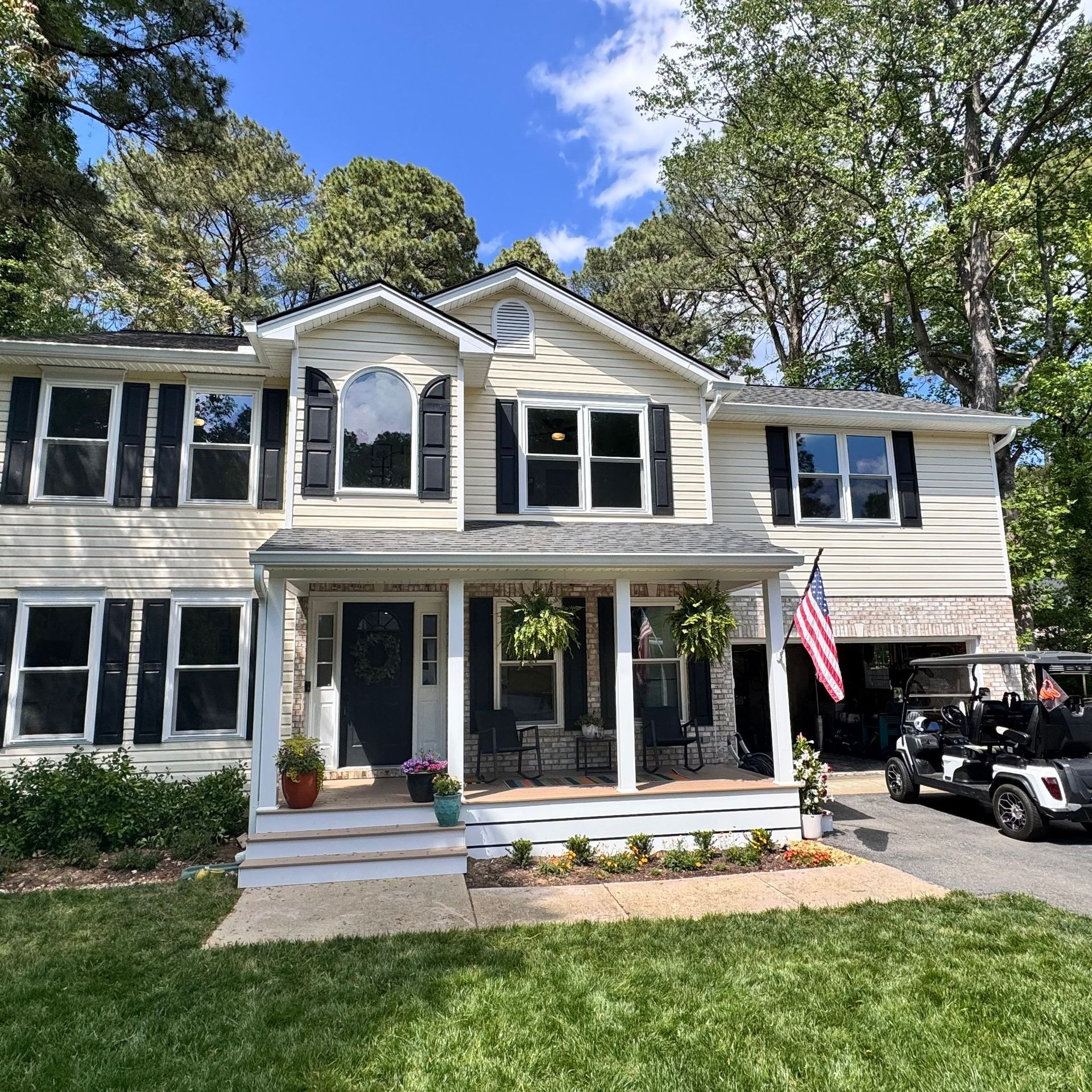 Two-story beige house with white trim, black shutters, front porch, and an American flag under a blue sky