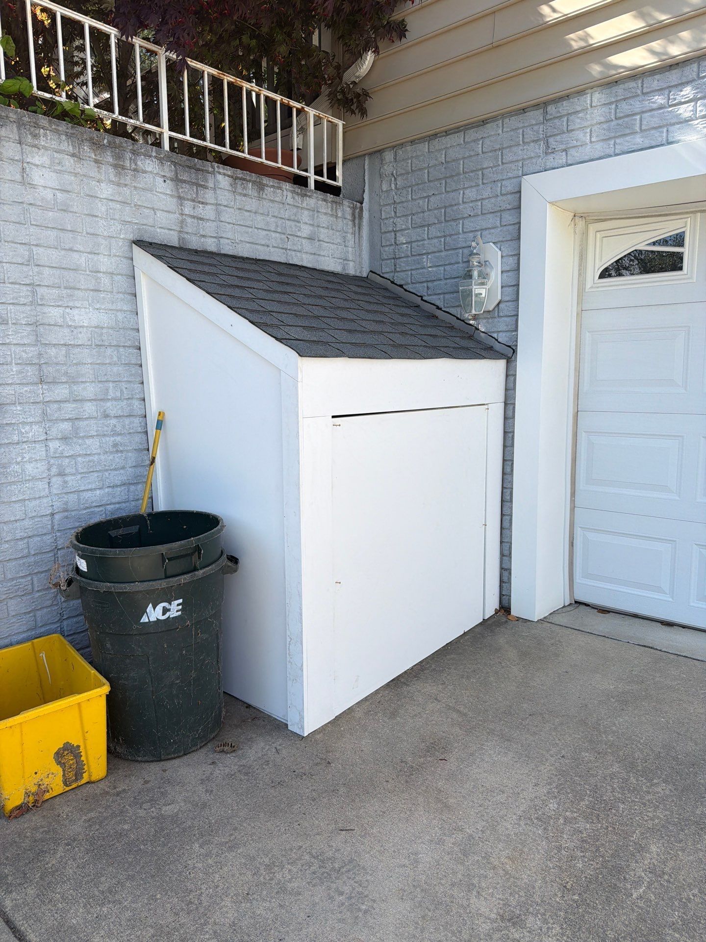 White storage shed beside a garage door on a gray brick patio, with a black trash bin and yellow container nearby.