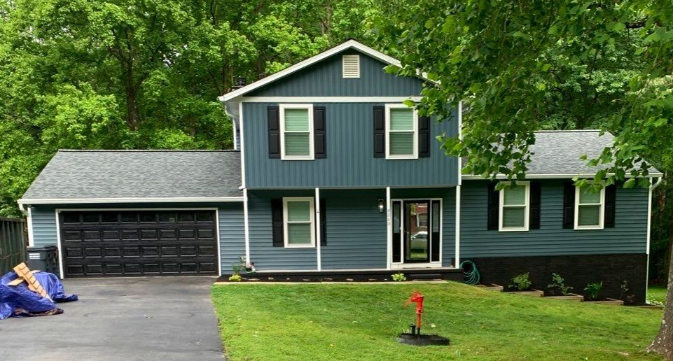 Blue two-story house with black garage and white trim on a green lawn, surrounded by trees
