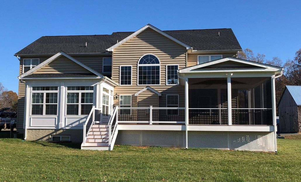 Two-story beige house with white trim, screened porch, and fenced yard under a clear blue sky.