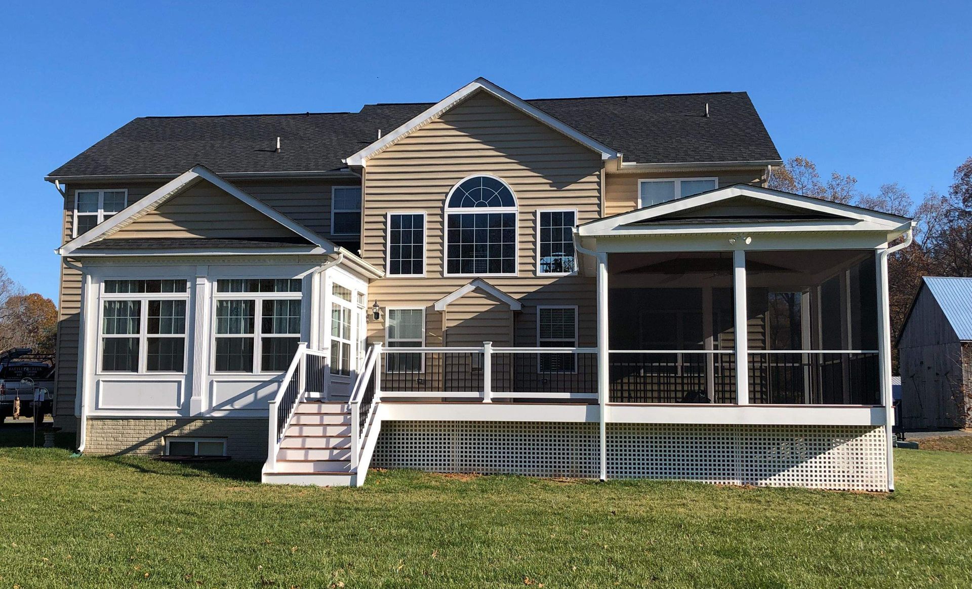 Two-story beige house with white trim, screened porch, and fenced yard under a clear blue sky.