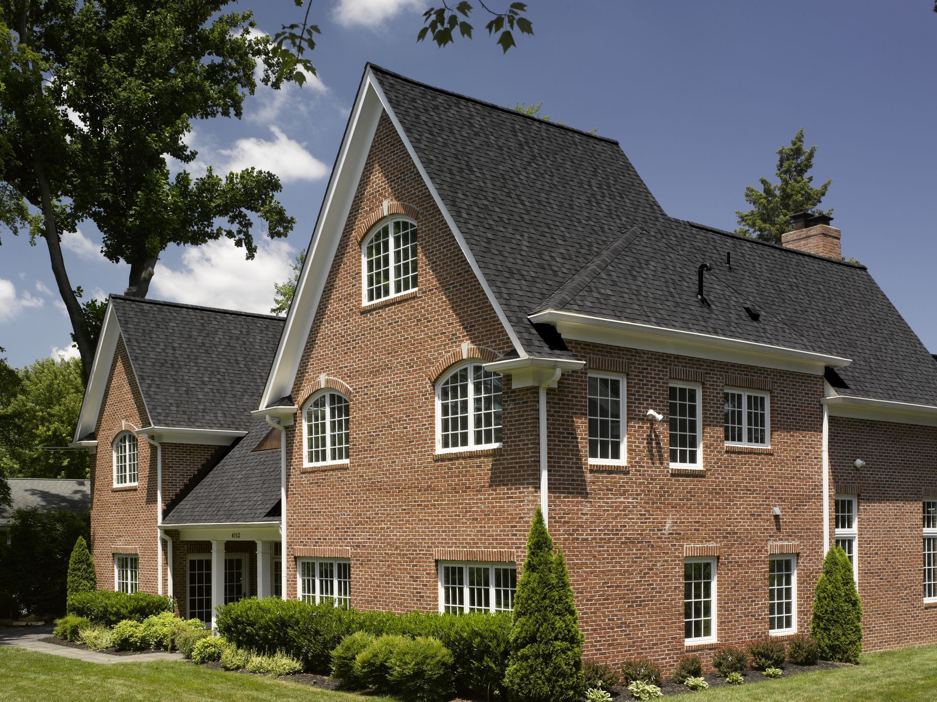 Large brick house with steep dark roof and white-trimmed windows, surrounded by green lawn and trees.