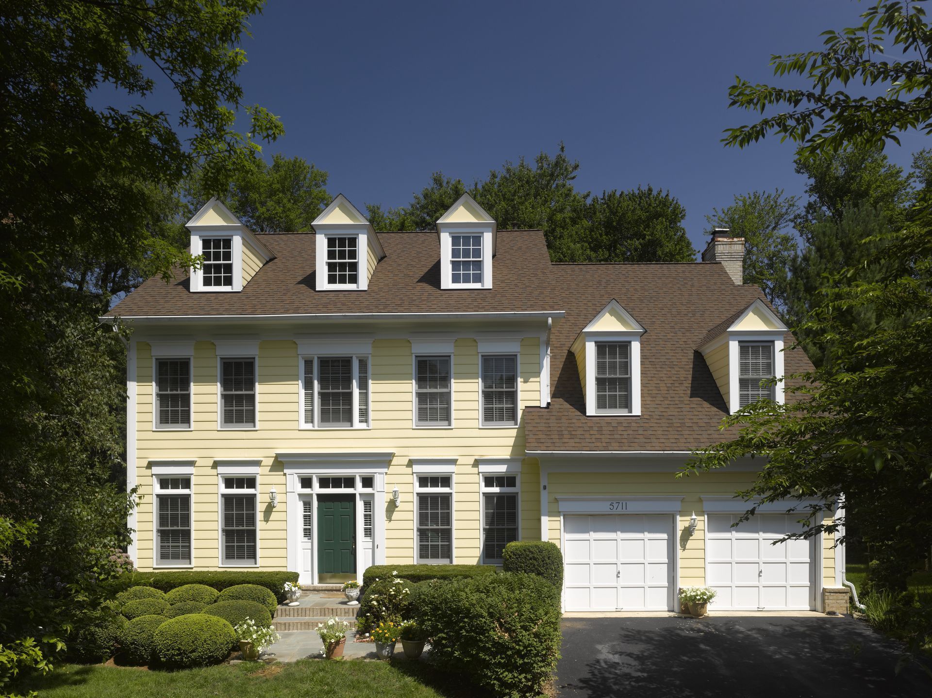 Large yellow suburban house with white trim, dormer windows, garage, and landscaped front yard under blue sky