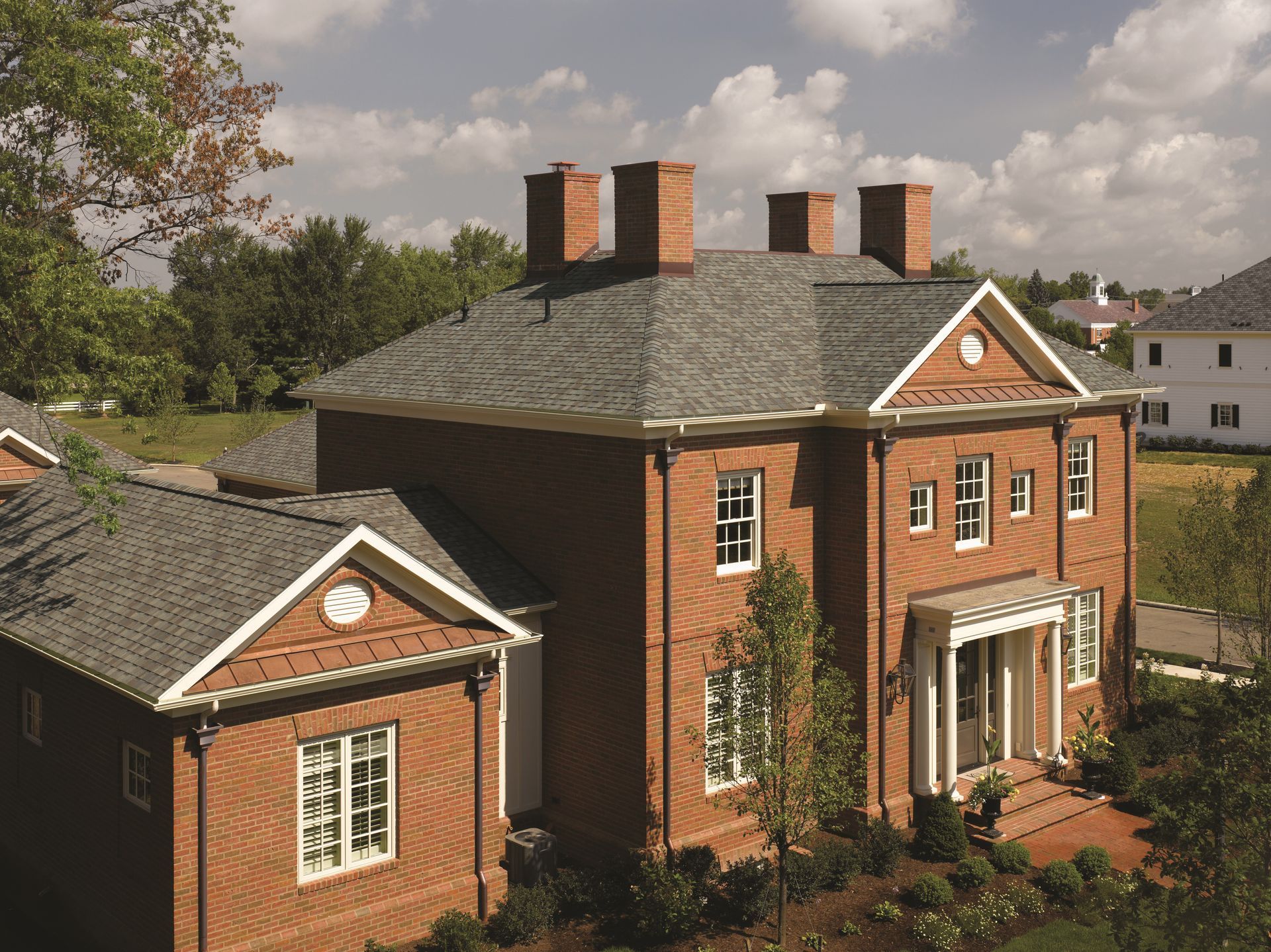 Large red-brick house with slate roof and chimneys, seen from above among trees and neighboring homes
