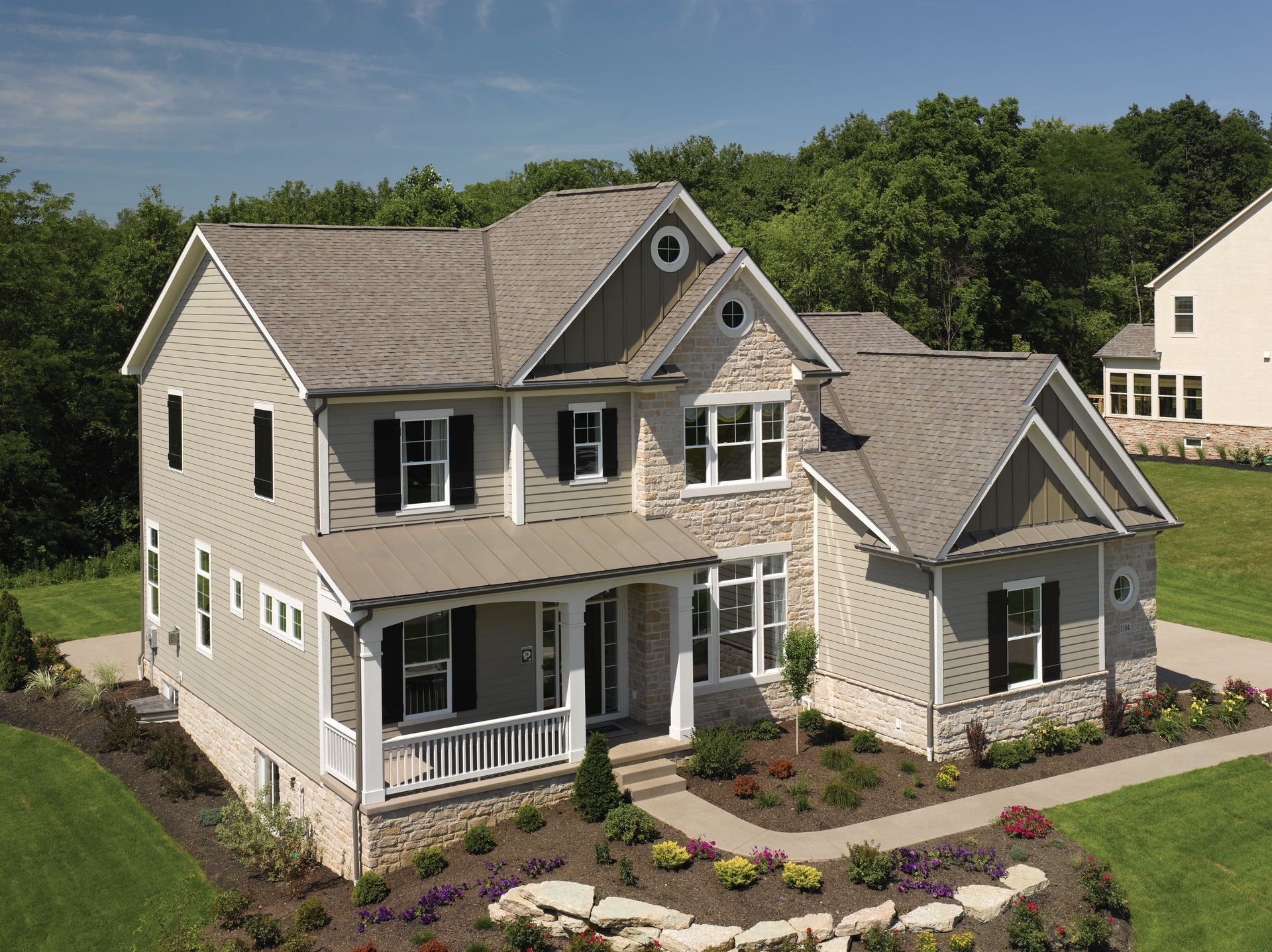 Suburban two-story beige house with front porch, dormer windows, and landscaped yard on a sunny day