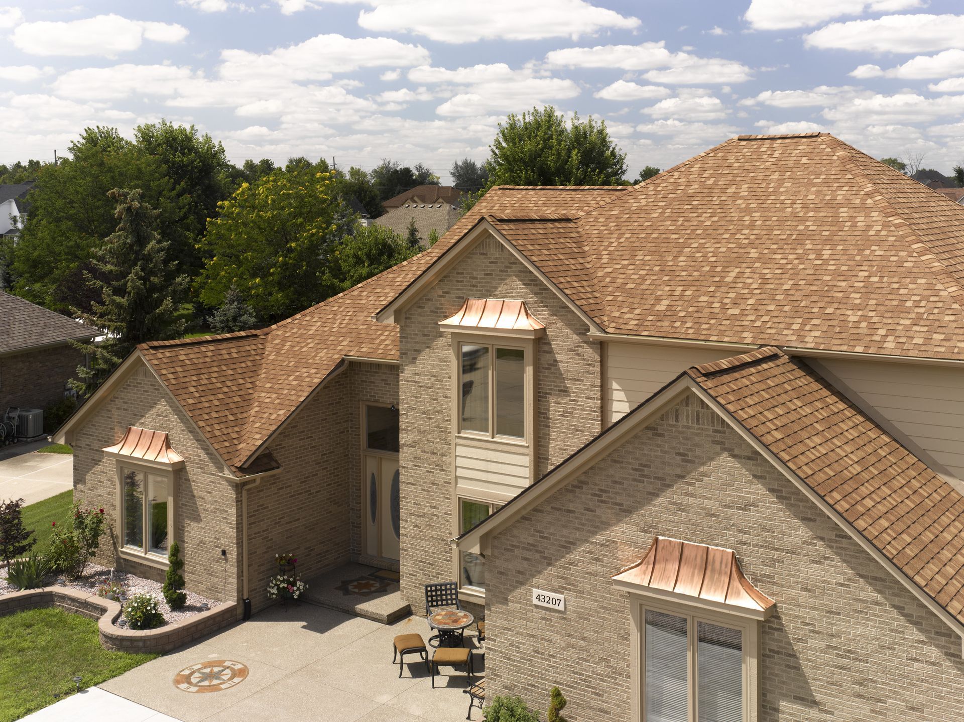 Stone house with tan shingle roofs, dormer windows, and a paved driveway surrounded by trees.