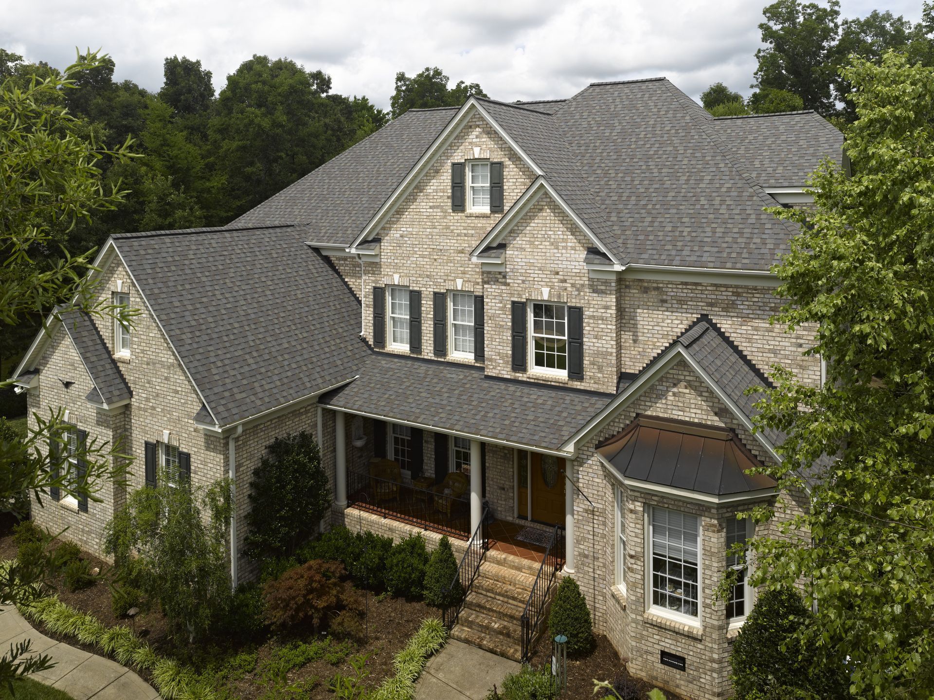 Large gray stone suburban house with a front porch and dark roof, surrounded by green trees and shrubs