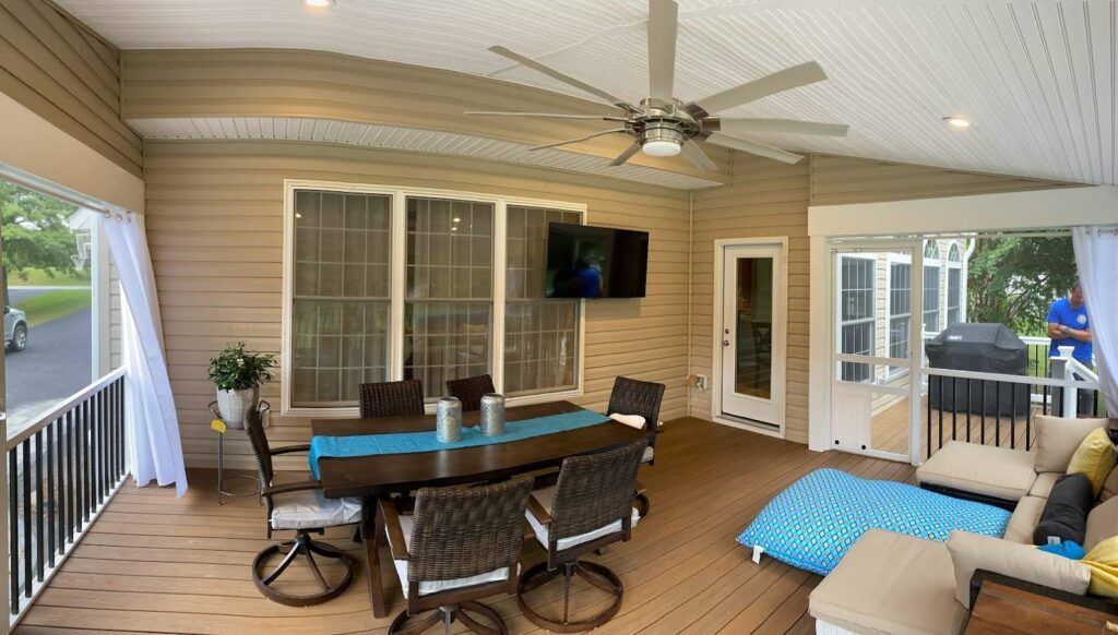 Covered patio with dining table, chairs, ceiling fan, TV, and blue lounge chairs beside sliding doors.