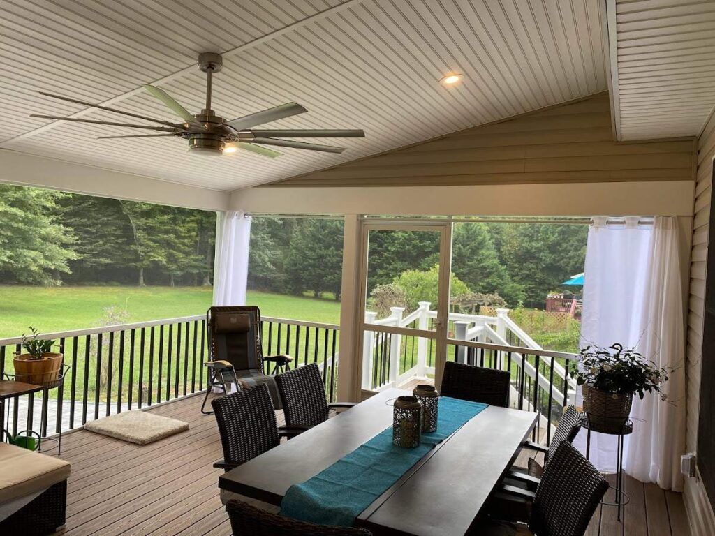 Covered porch with dining table, ceiling fan, grill, and view of a green backyard through white curtains.