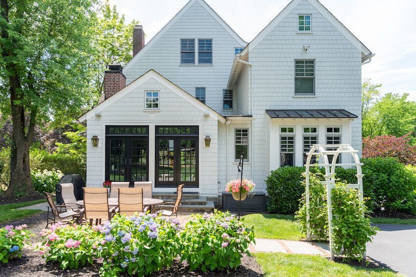 White two-story house with patio seating, flower garden, and arched trellis in a sunny yard