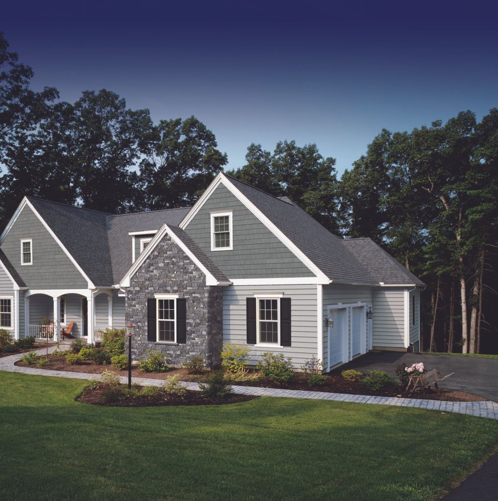 Gray suburban house with white trim, stone facade, manicured lawn, and trees at dusk
