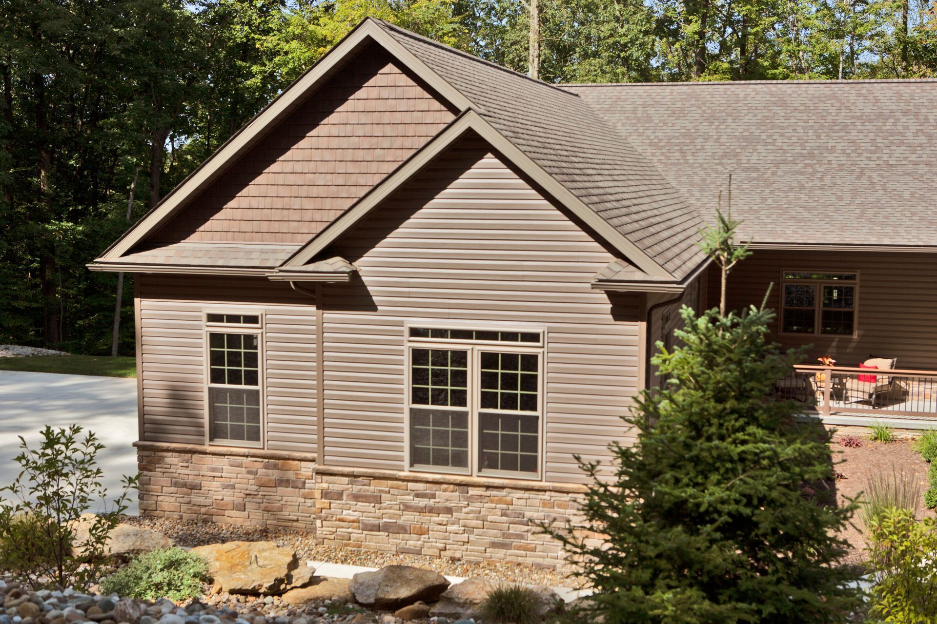 Beige house with stone foundation, gray roof, and large windows amid trees and landscaping