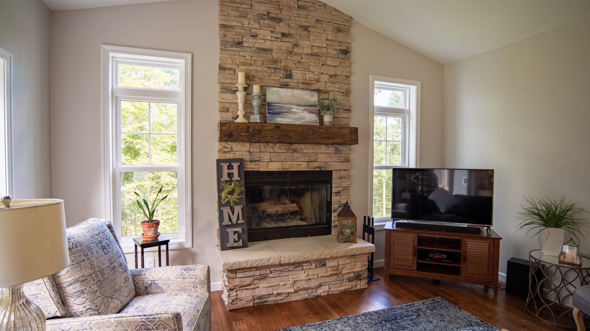 Bright living room with a stone fireplace, tall windows, armchair, TV, and hardwood floors.