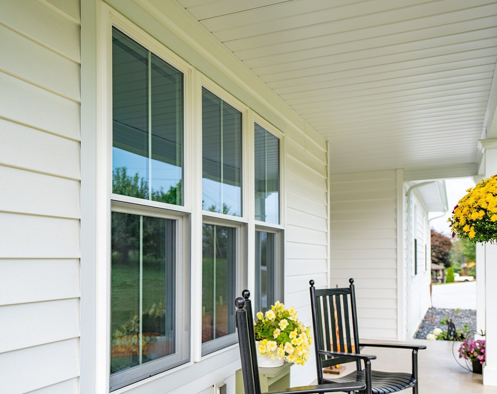 White porch with windows, black chairs, and potted yellow flowers along a bright covered walkway.