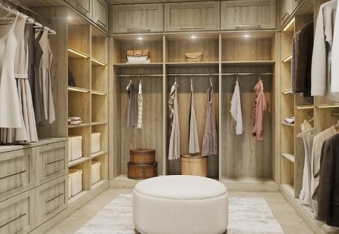 A modern, well-organized walk-in closet with light wood cabinetry, hanging clothes, shelves, and a white ottoman.
