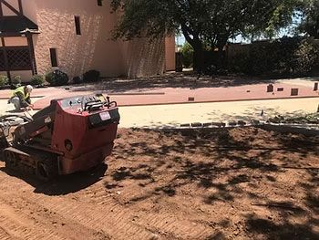 Red construction machine on bare dirt beside a house and pink sidewalk in a sunny yard