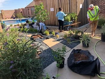 People landscaping a backyard with rocks, plants, and a hose beside a wooden fence and pool.