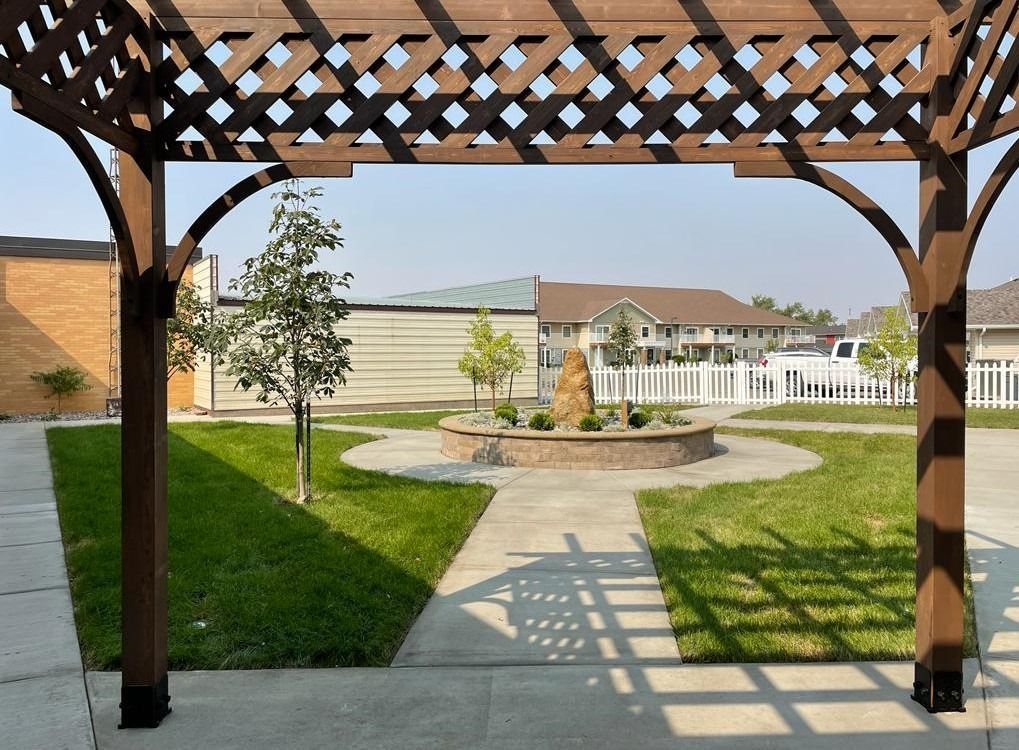 Covered garden walkway leading to a patio, with a lawn, young trees, and a white pergola in the background