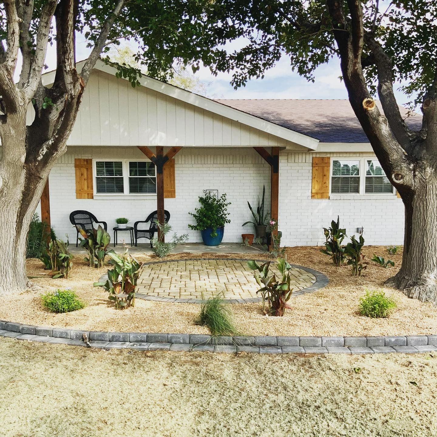 White house with front porch, potted plants, two chairs, and a gravel yard shaded by large trees