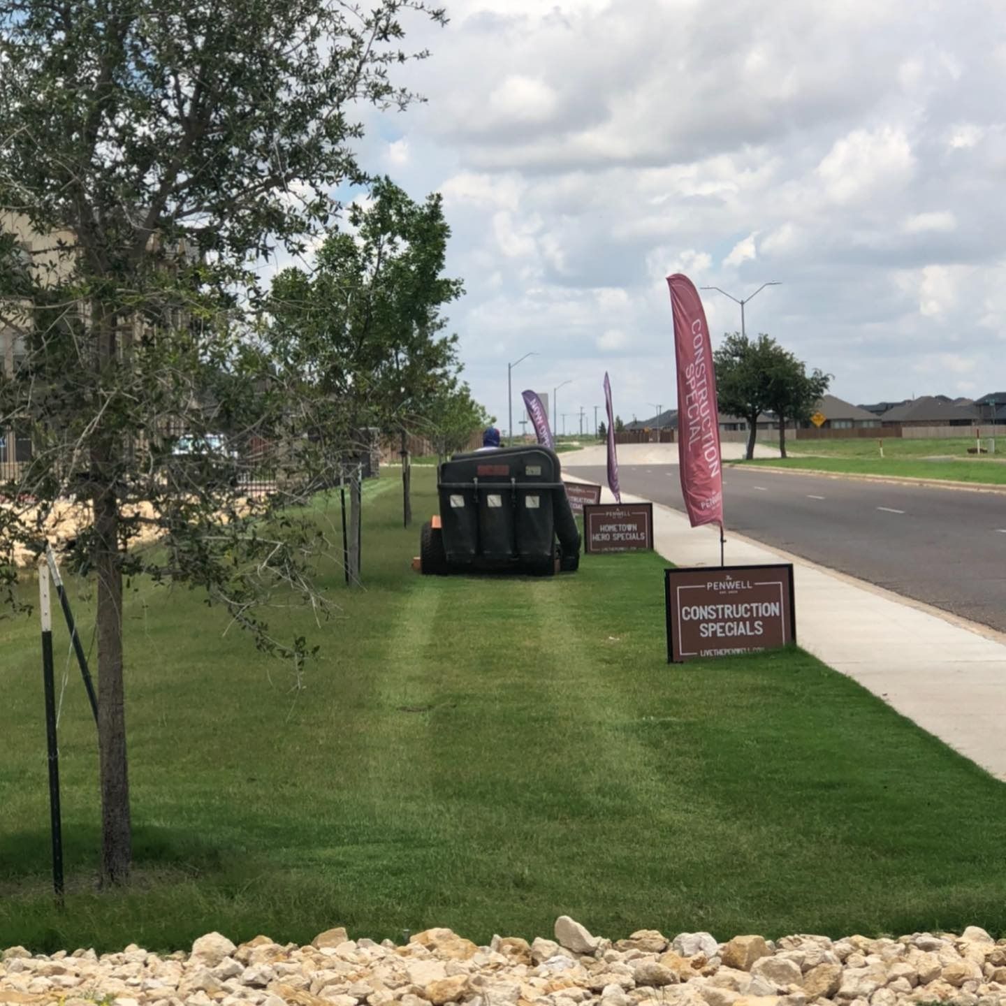 Grassy roadside with a black utility cart, trees, and a sign near a paved path under cloudy skies