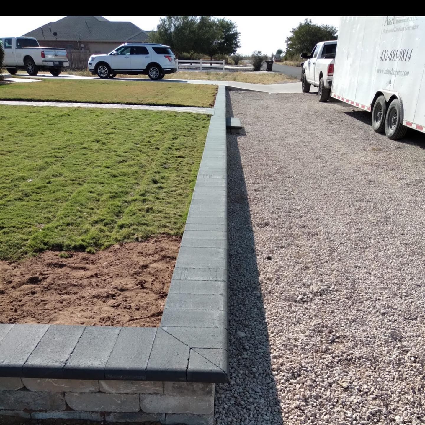 Gravel driveway beside a raised lawn bordered by gray pavers, with parked vehicles in a residential area.
