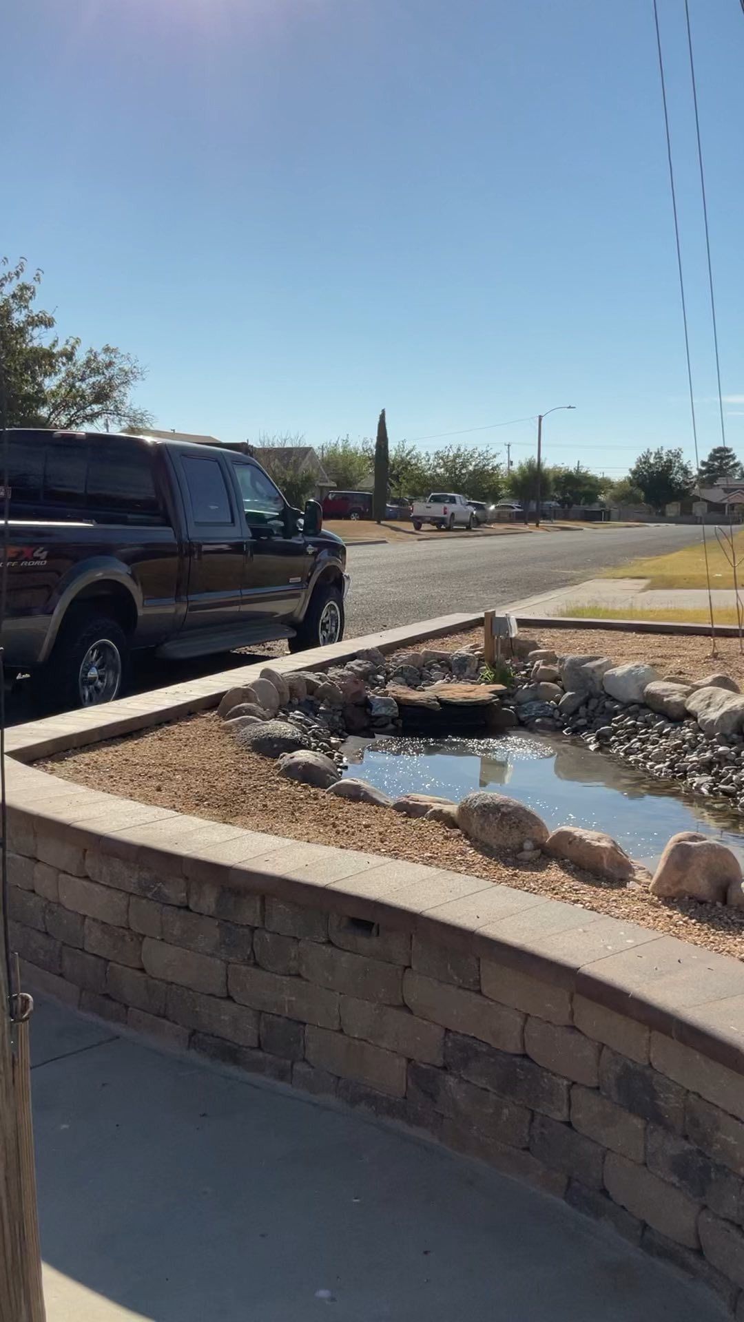 Landscaped roadside fountain with rocks and water beside a parked black SUV under a clear blue sky