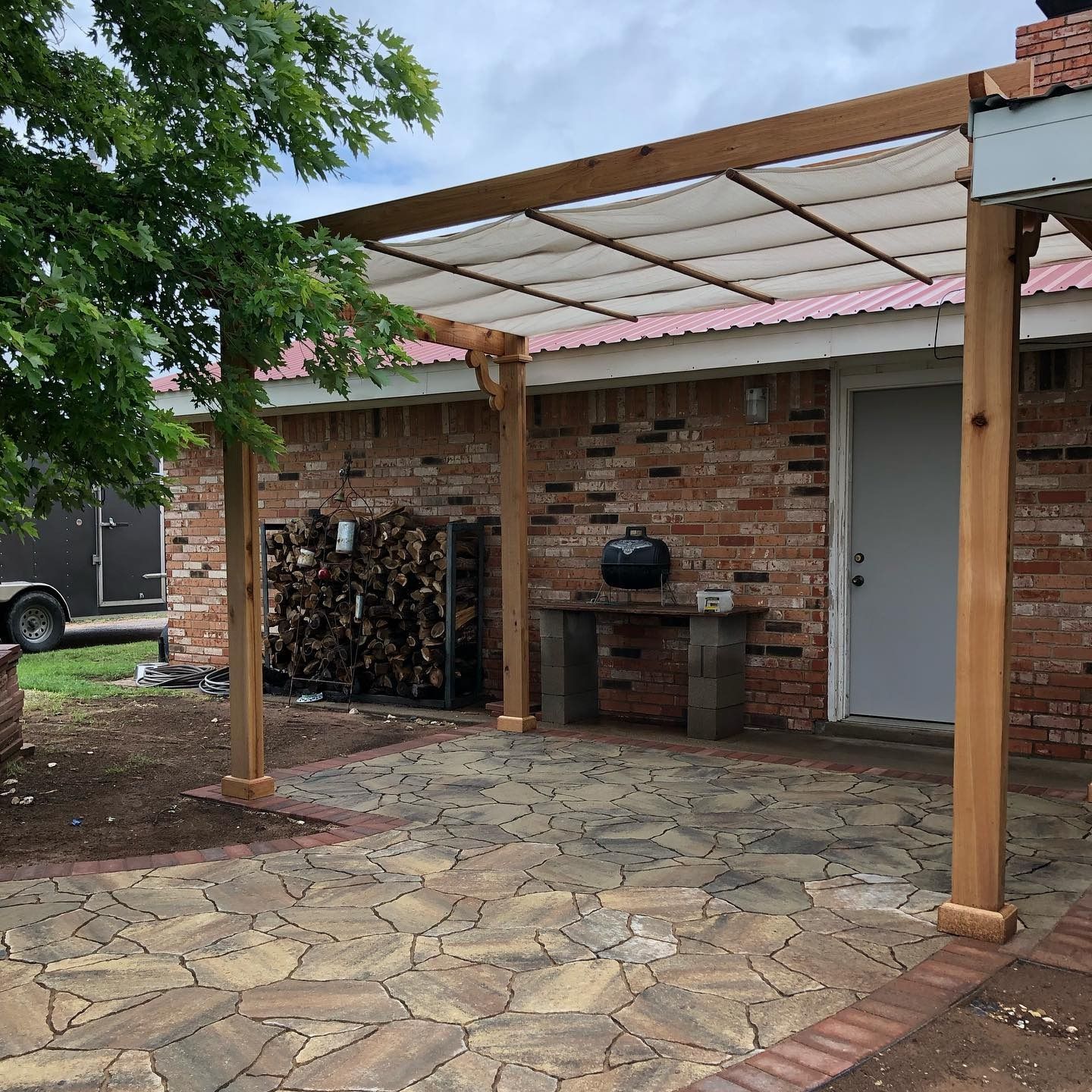 Brick patio under a wooden pergola beside a brick house with a white door and grill.